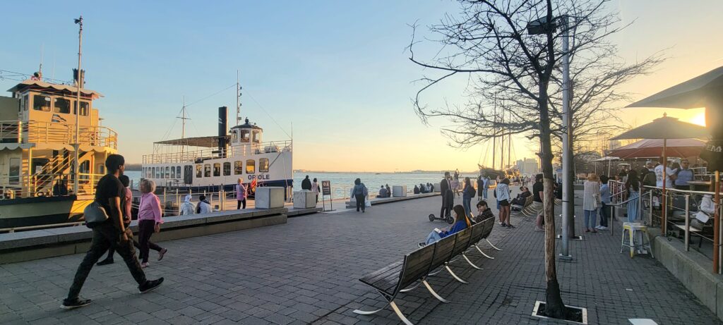 Boats, restaurants, and people along the Harbourfront, free to visit in Toronto, shown with the sun setting in the background.
