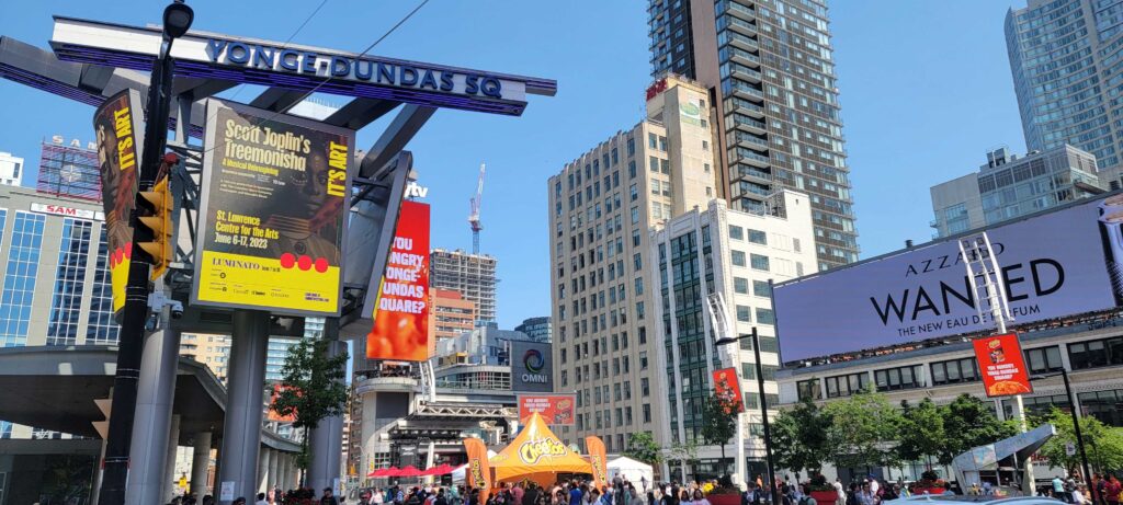 Yonge Dundas Square on a sunny afternoon, free to visit in Toronto, shown with crowds of people, signs, and buildings.