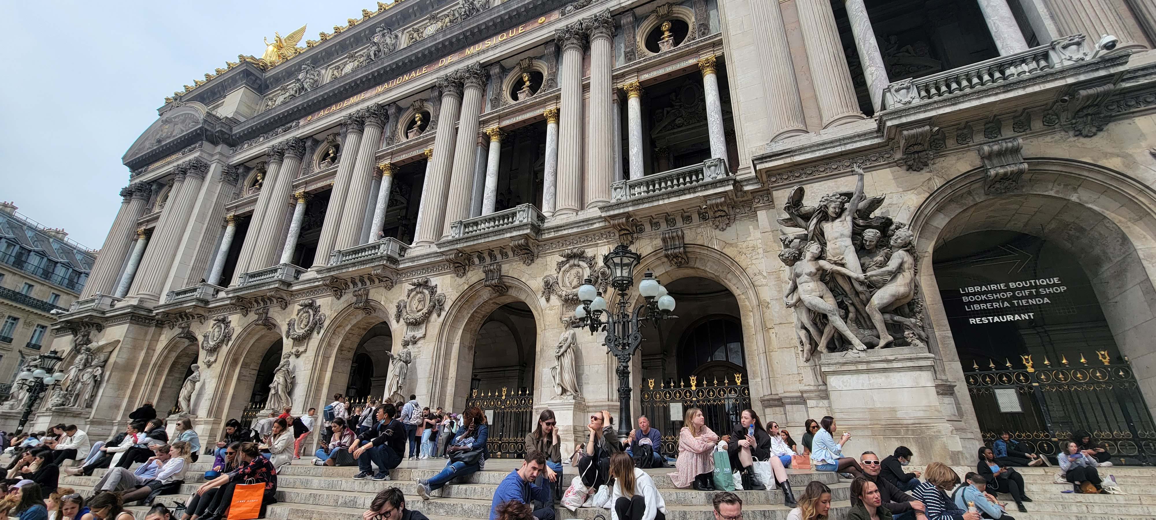 Exterior of Palais Garnier, Paris France with people sitting on the steps outside.