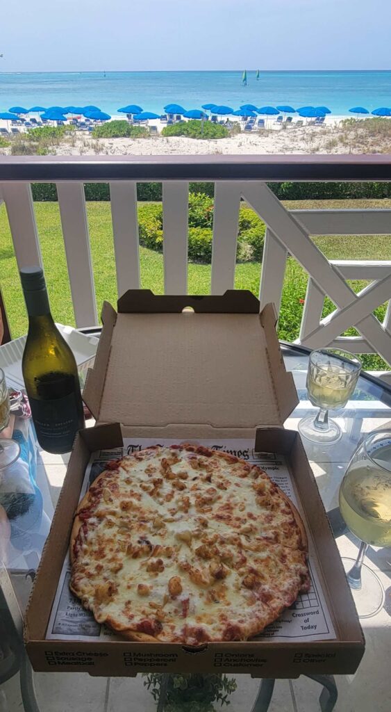 Take-out pizza and bottle of wine on table on balcony with beach and ocean view in the background.