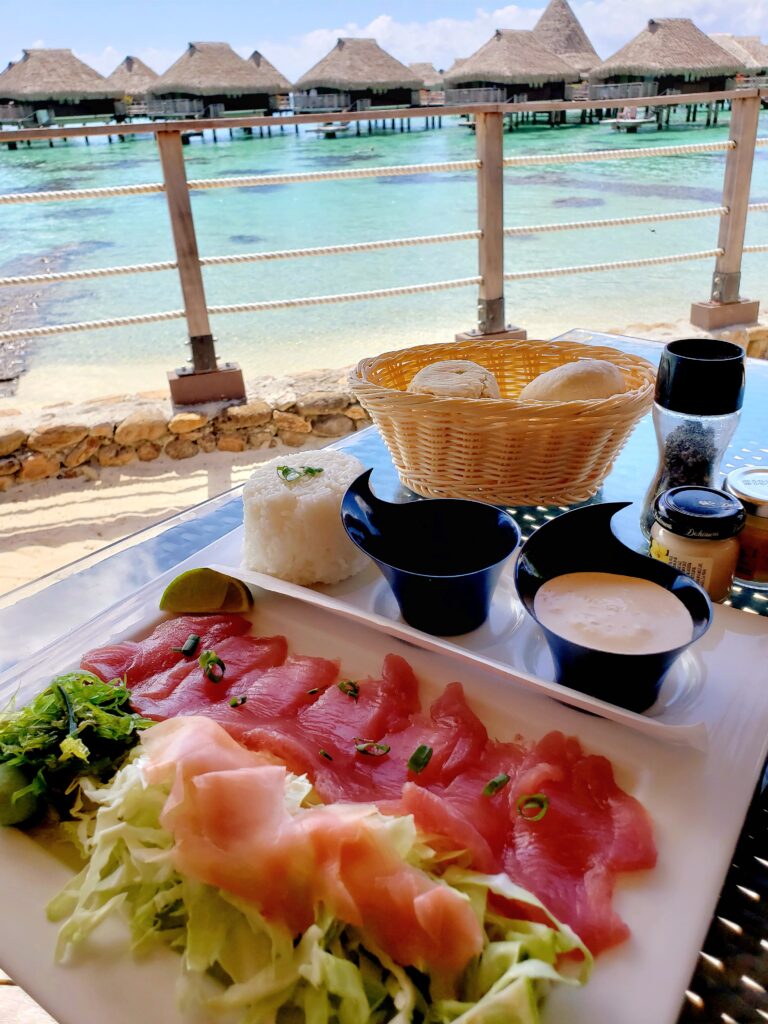 Sashimi, salad, bread basket ordered for lunch with overwater bungalows in the background at the Hilton Moorea Lagoon Resort & Spa,