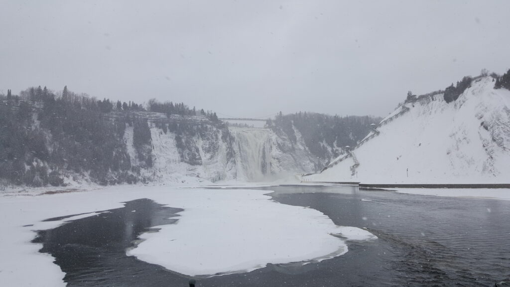 Montmorency Falls on a winter day at the base of the falls with snow on the surrounding hills and water.