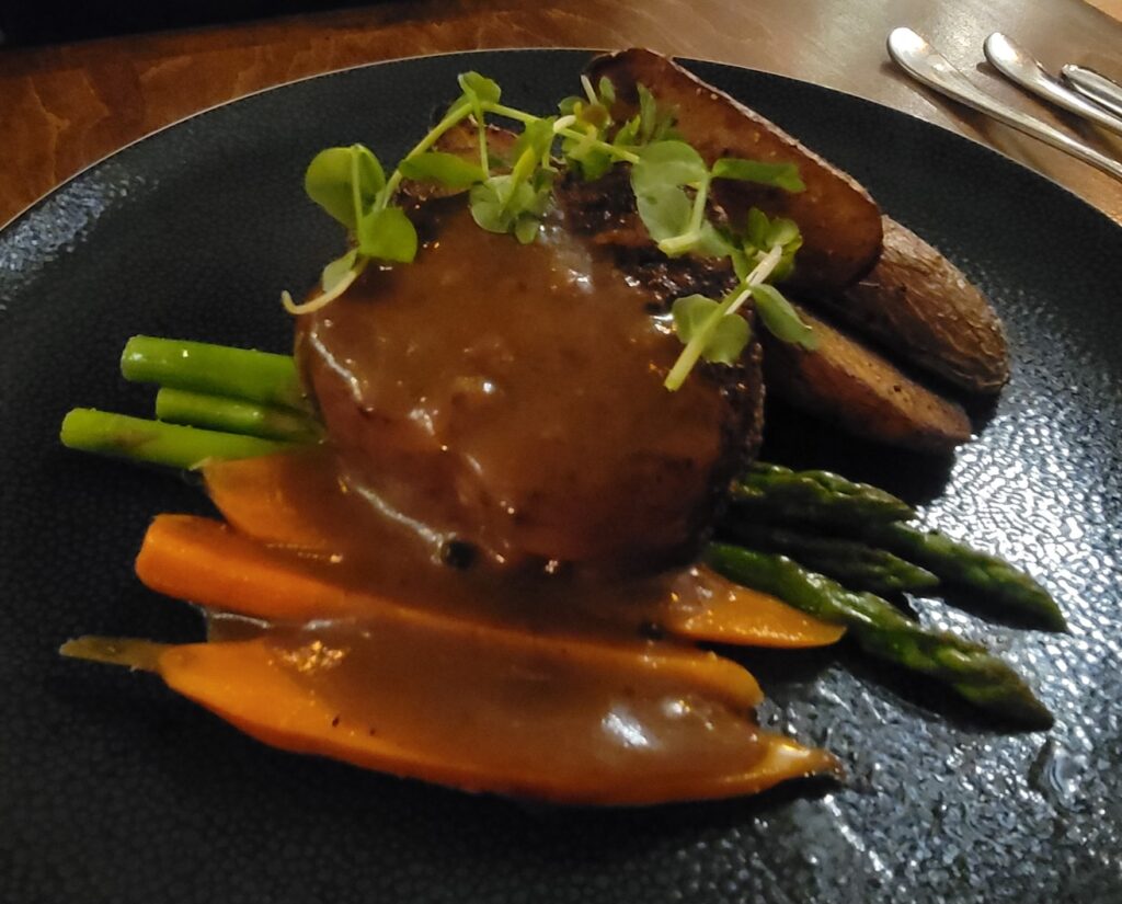 Close up of beef tenderloin with peppercorn sauce, served on a black plate, with potato, asparagus, carrots from Zees Grill diner menu.