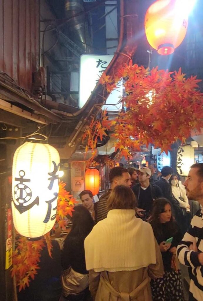 Walking through crowded "Memory Lane", Omoide Yokocho, with lit paper lanterns and fall leaves on a busy evening.