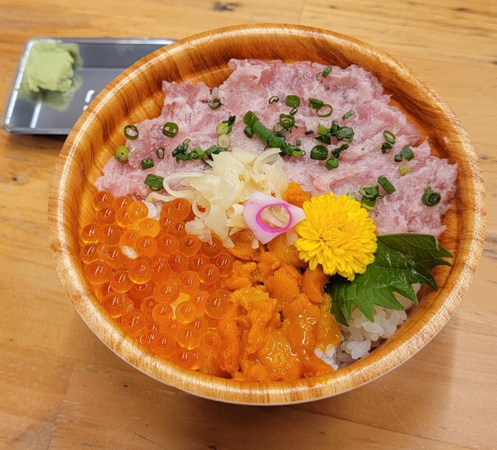 Close up of sea urchin, salmon roe & minced fatty tuna on rice bowl from Tsukiji Outer Market, with ginger and yellow flower on top.