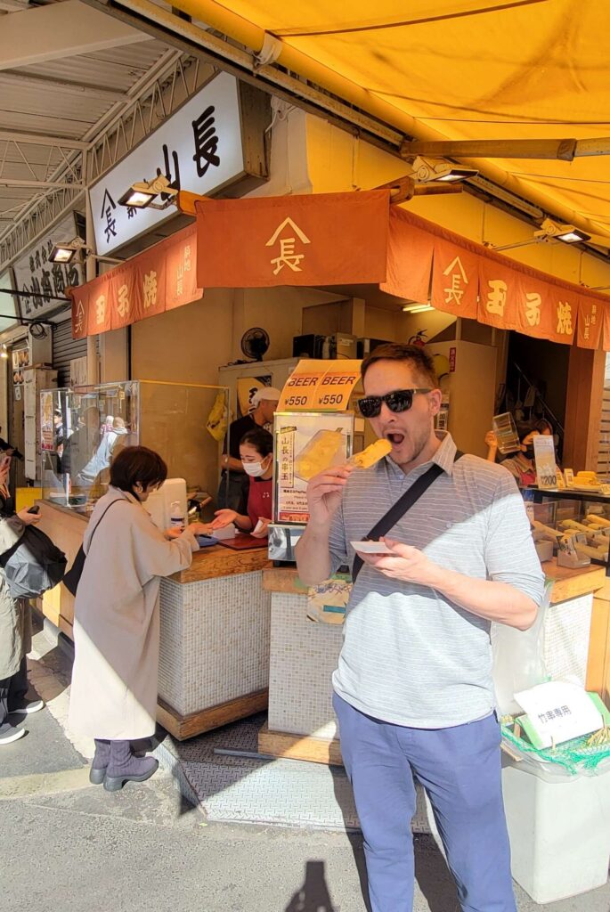 Man with sunglasses eating tamago yaki in front of food stall at Tsukiji Outer Market.