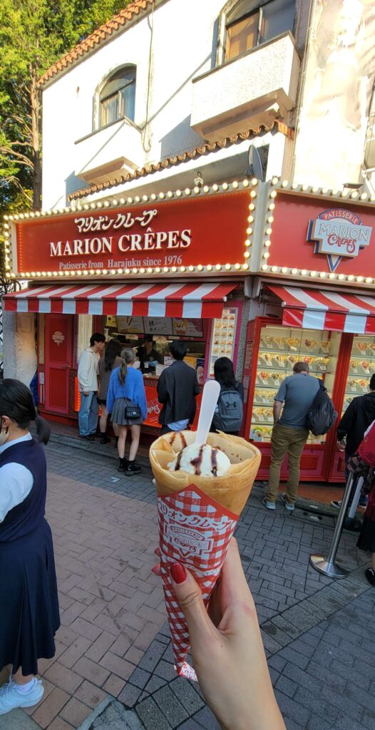 Female hand holding a crepe from Marion Crepes, with the store and people lined up in the background, on Takeshita Street.