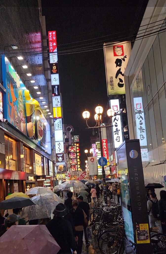 Crowds walking around the bright lights and restaurants in Osaka, many with umbrellas, despite the rain, at night.