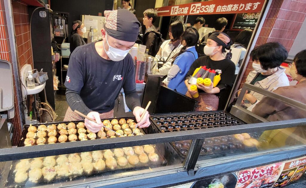 Takoyaki being cooked outside in Osaka with people waiting in line to order.