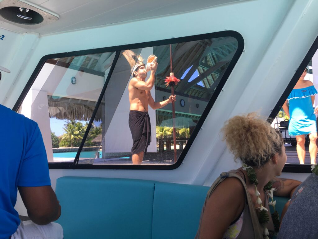 Window view from inside the boat upon arrival at the InterContinental Bora Bora & Thalasso Spa, of man dressed in traditional Tahitian attire, welcoming us with conch shell blowing.