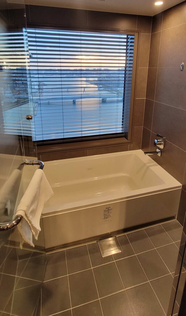 Glass door opened to show separate bathing room with shower, white tub, and window with a view of Tokyo Bay, at Mitsui Garden Hotel Toyosu Premier.