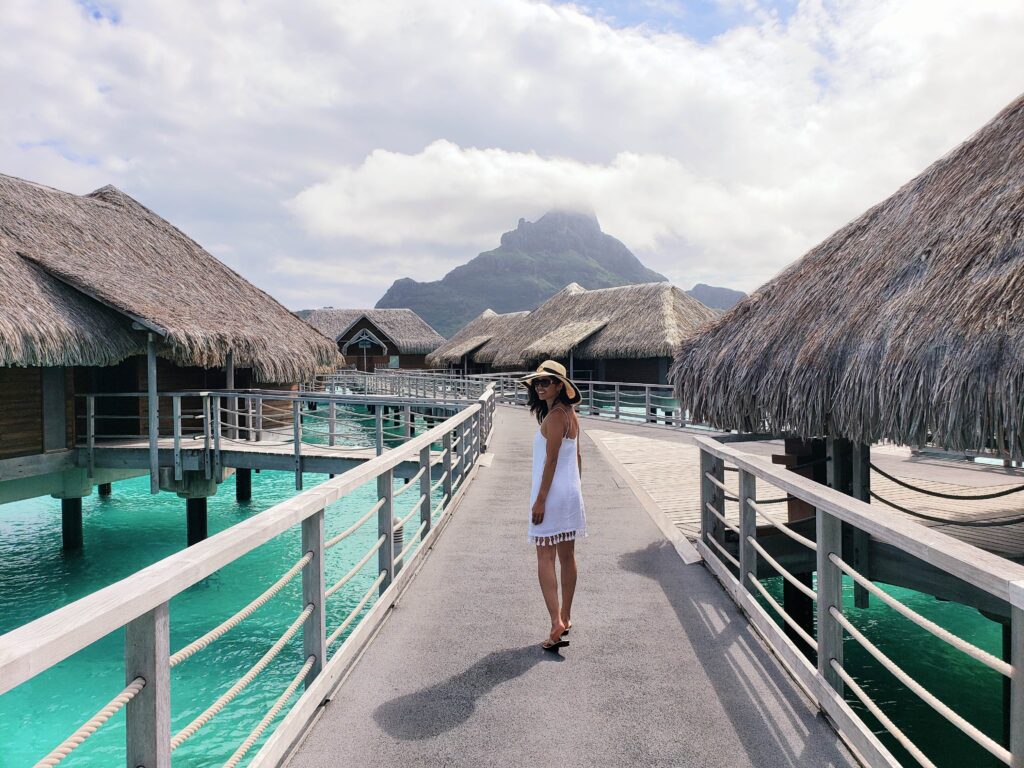 Woman with sun hat and white dress looking back smiling, walking by the overwater villas at InterContinental Bora Bora & Thalasso Spa.