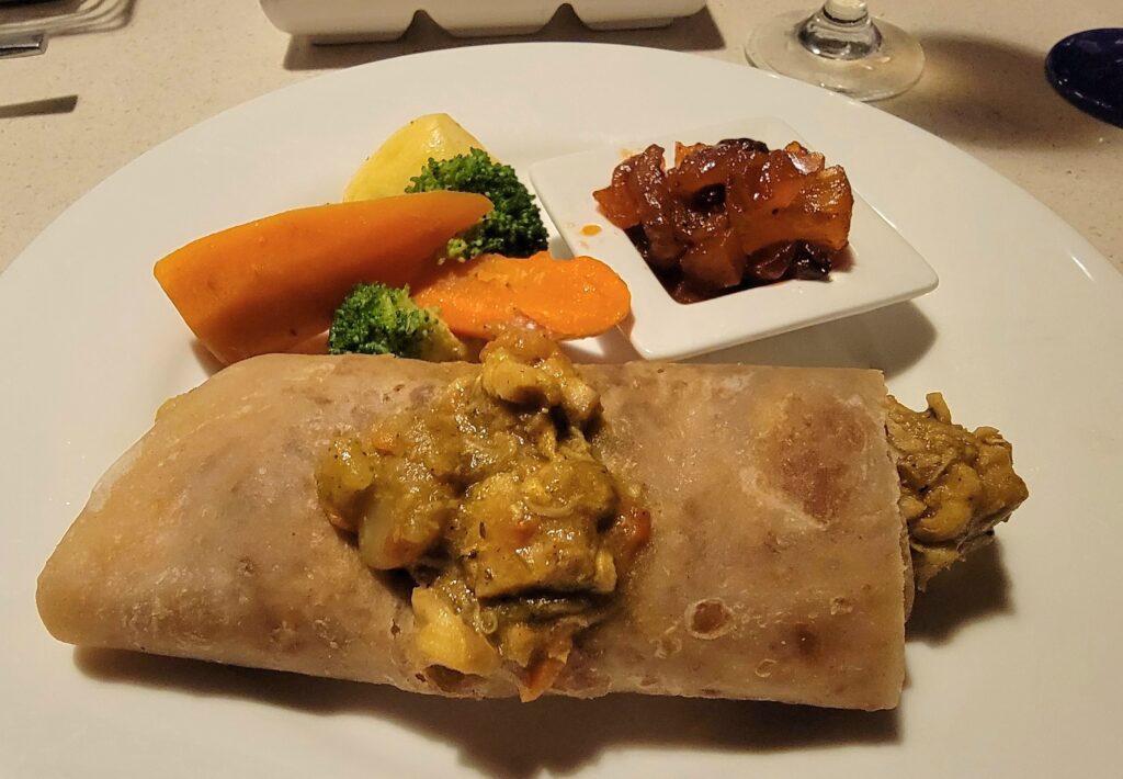 Roti filled with curry chicken, from Sandals Grenada, on a white plate served with a small side of vegetables and fruit chutney.