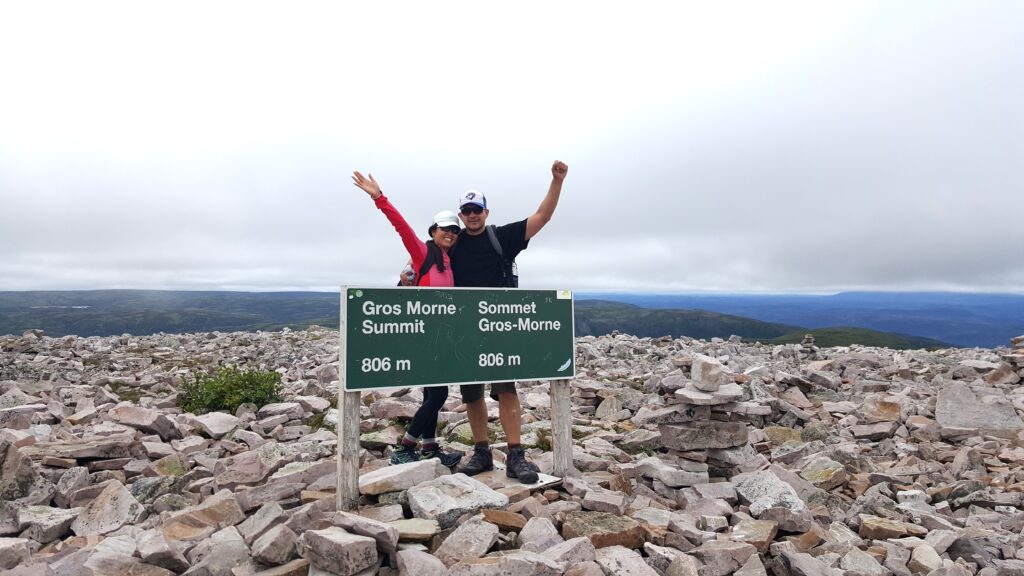 Happy couple with arms up at the Gros Morne Summit mountain sign in Newfoundland, a great trail for forest bathing and nature.