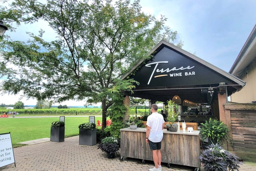 Man in black shorts and white shirt standing at the entrance of Peller Estates Terrace Wine Bar on a sunny day.