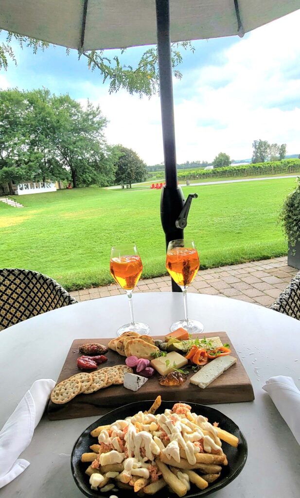 Ontario cheese board, lobster poutine and two spritz at Peller Estates Terrace Wine Bar, with Peller sign and vineyards in the background, on a sunny day.